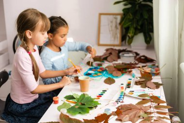 Kızlar sonbahar yapraklarını boyarlar. Gouache, fırça ve ahşap bir masada çeşitli sonbahar yaprakları. Çocuk sanat projesi. Kuru sonbahar yapraklarında renkli elle boyanmış.