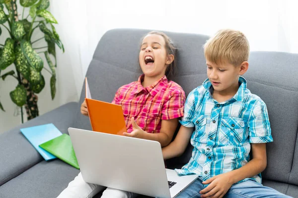Niño y niña, hermano y hermana estudian en casa. Una chica ve una lección de video o una conferencia en línea, un niño hace ejercicios en un cuaderno de un libro de texto — Foto de Stock
