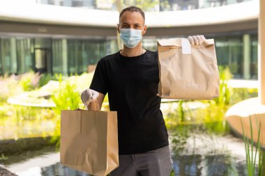 COVID-19 pandemik tecrit konsepti sırasında temassız teslimat. Maske ve eldiven takan kurye bir paket kutusu tutuyor ve akıllı telefondan adres bilgilerini kontrol ediyor.