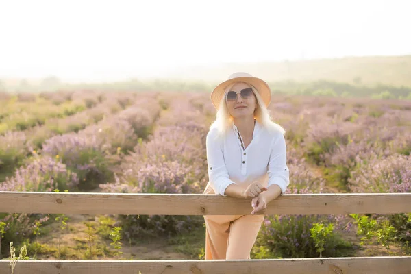 Jovem mulher loira viajante usando chapéu de palha no campo de lavanda cercada com flores de lavanda. — Fotografia de Stock