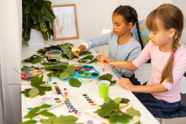 Kızlar sonbahar yapraklarını boyarlar. Gouache, fırça ve ahşap bir masada çeşitli sonbahar yaprakları. Çocuk sanat projesi. Kuru sonbahar yapraklarında renkli elle boyanmış.