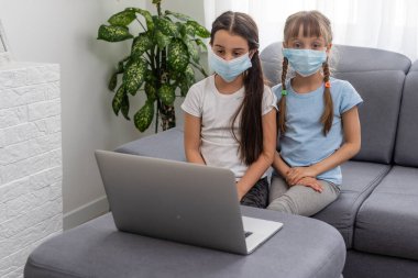 Mutlu iki küçük kız öğreniyor, çevrimiçi çalışıyor ve yüz maskesi ile dizüstü bilgisayarda çalışıyor.