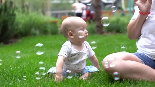 Junge sitzt auf grünem Gras im Freien und spielt mit Seifenblasen — Stockvideo