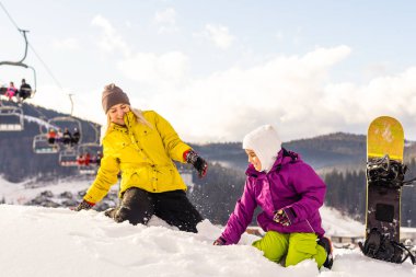 Snowboard yapan anne ve kızı karda oynuyorlar.