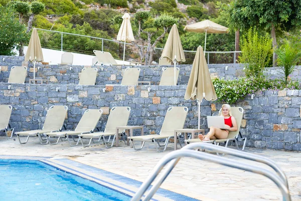 Vista frontal de la mujer happ utilizando el ordenador portátil mientras se relaja en una tumbona en el patio trasero en casa. Diversión de verano en casa junto a la piscina — Foto de Stock