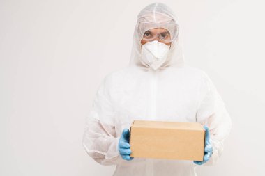 Beyaz arka planda elinde bir paketle tehlikeli madde giysisi içinde duran bir adam.
