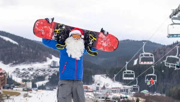 Papai Noel com snowboard contra neve inverno montanha esqui resort paisagem e céu azul, Ano Novo ou xmas cartão. — Fotografia de Stock