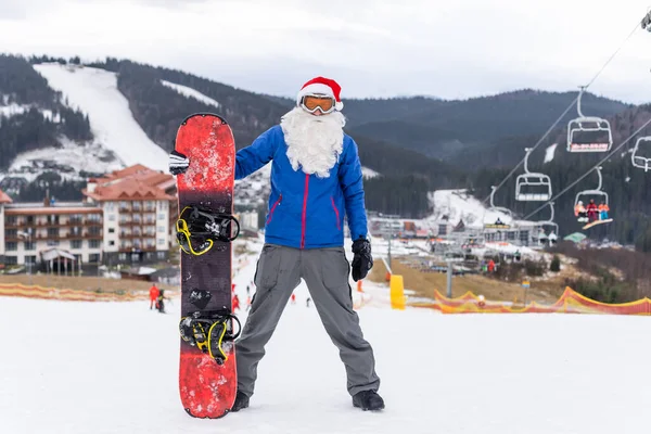 Glücklicher Mann mit santa-roter Weihnachtsmütze und Snowboard am Winterberg — Stockfoto