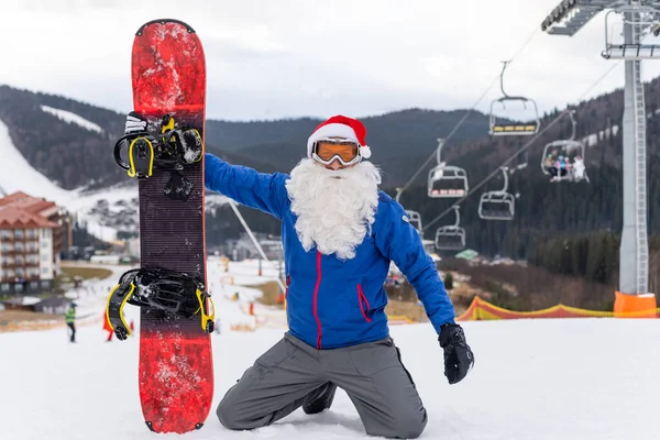 Egy férfi télapó kalapban, snowboarddal a síközpontban. — Stock Fotó