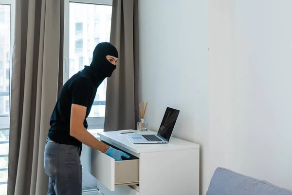 Κλέφτης με μαύρη κουκούλα που κλέβει λάπτοπ. Ο διαρρήκτης διαπράττει έγκλημα σε πολυτελές διαμέρισμα — Φωτογραφία Αρχείου