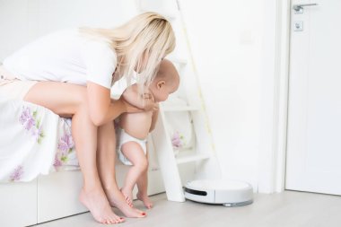 Genç anne yeni yürümeye başlayan oğluyla kanepede oturuyor ve robot elektrik süpürgesinin ev işi yapmasını izliyor..