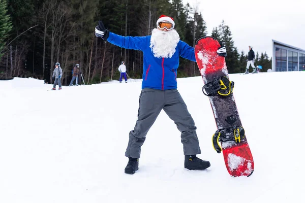 Kerstman met snowboard tegen besneeuwde winter bergskigebied landschap en blauwe lucht, Nieuwjaar of kerst wenskaart. — Stockfoto