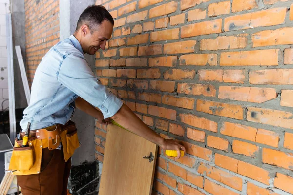 Молодий професіонал у порожній незакінченій квартирі вимірює стіну за допомогою мірної стрічки, перевіряючи ноти. Концепція ремонту та ремонту — стокове фото