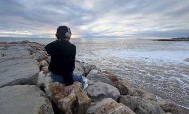 Kız plajı keşfediyor, kulaklık takıyor, okyanusa bakıyor, sakin bir ifadeyle ve bulutlu bir arkaplanla, gezinti şehvetini yakalıyor. Yüksek kalite fotoğraf