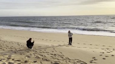 Sahilde, gün batımında deniz kıyısında iki yolcunun mobil fotoğraf çekimi. Sahilde poz veren genç bir kadın ve onu çeken bir kız. Ağır çekim, arka ışıklandırma, yerel turizm. Yüksek kalite 4k görüntü