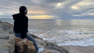 Sarı ceketli genç kız bir kayanın üzerinde oturuyor ve okyanus ve kayalıklarla büyüleyici doğa geçmişine bakıyor. Seyahat ve turizm. Portekiz. Dramatik gün batımı gökyüzü. Meşhur bir bölge. Yüksek kalite fotoğraf