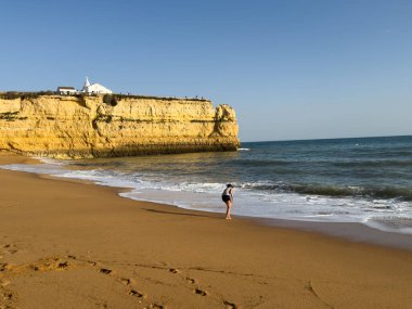 Günbatımında sakin bir plajın sığ dalgalarında, arka planda uzak bir uçurum bulunan tek bir kişi duruyor. Sahne yalnızlığı, sakinliği ve doğayla bağlantıyı çağrıştırıyor. Yüksek kalite fotoğraf
