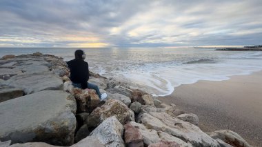 Kapüşonlu ve bereli biri Portekiz sahilinde sakin bir gün batımını seyrediyor. Yüksek kalite fotoğraf