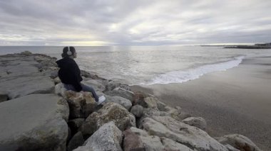 Sarı ceketli genç kız bir kayanın üzerinde oturuyor ve okyanus ve kayalıklarla büyüleyici doğa geçmişine bakıyor. Seyahat ve turizm. Portekiz. Dramatik gün batımı gökyüzü. Meşhur bir bölge. Yüksek kalite fotoğraf