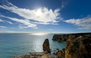 Karanlık kayalıklı dramatik bir deniz manzarası. Altın güneş ışığı dramatik fırtına bulutlarını yararak parlak turuncu ve sarı röfleler oluşturur. Yüksek kalite fotoğraf