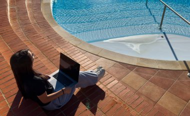 Yüzme havuzunun yakınında dizüstü bilgisayar kullanan serbest çalışan biri. Genç gezgin kız yaz tatilinde bilgisayarda çalışıyor. Teknoloji ve yaşam tarzı konsepti, kopyalama alanı. Yüksek kalite fotoğraf