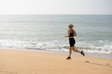 Kumsalda koşan bir kadın, okyanus manzarasının tadını çıkarıyor. Kumsal koşusu, okyanus esintisi ve fitness işin püf noktası. Deniz kenarında aktif bir yaşam tarzı, plaj atmosferini kucaklamak. Kumsalda egzersiz yapan bir kadın. Yüksek kalite fotoğraf