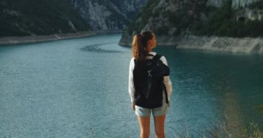 Büyük sırt çantalı mutlu bir turist dağın tepesinde duruyor ve dağ gölü ve dağların güzel manzarasını düşünüyor. Günbatımı akşamı. Arka plan