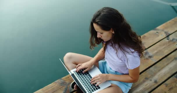 Een jonge brunette zit achter een houten pier bij het meer en typt tekst op een laptop. Zicht van bovenaf. Uitzicht op de houten pier en het meer — Stockvideo