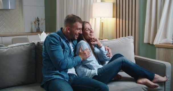 Um casal passa tempo sentado no sofá, abraçando e se divertindo conversando, assistindo TV. Retrato feminino e masculino — Vídeo de Stock