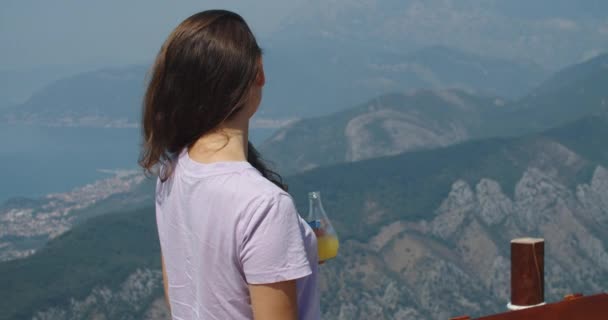 Jovem morena feliz fica nas montanhas em uma altura, bebe suco de laranja, aproveitando o momento. Retrato feminino. Vista montanha. — Vídeo de Stock
