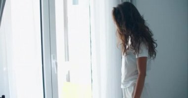 Günaydın Uyan. Çekici, gülümseyen genç bir kadın uyandığında tamamen beyaz bir yatakta dinleniyor. Yataktan kalkıp büyük pencereye doğru yürürken kollarını uzatan güzel, sağlıklı bir manken kız..