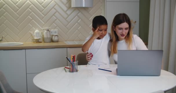 Beyaz anne ve küçük Afrikalı oğlu evdeki mutfakta oturuyorlar. Bir anne dizüstü bilgisayarda çalışır, küçük yaramaz oğlu onun dikkatini dağıtır. Annem çalışırken oğlunu resim yapmaya davet ediyor.. — Stok video
