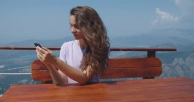Kıvırcık saçlı mutlu kadın turist bir kafede yükseklerde oturuyor. Cep telefonundan mesaj okuyor. Yükseklerden dağlara ve koylara bak..