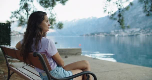 Mujer feliz se sienta en un banco de madera y considera hermosas vistas del lago y las montañas — Vídeo de stock