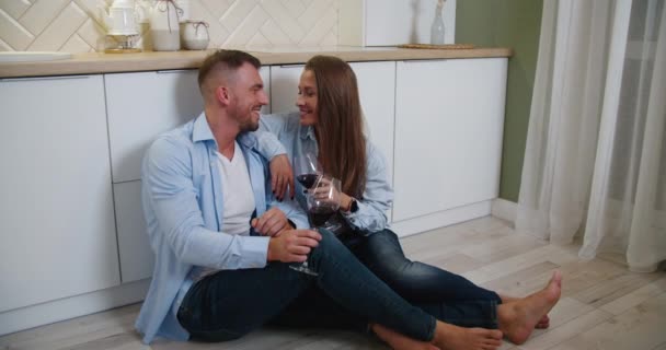 A fiatal szerelmesek vörösbort iszogatnak a konyhában, a padlón ülve. Boldog Y-generációs emberek otthon élvezik aperitif időt éljenzés együtt jubileumi évfordulója — Stock videók