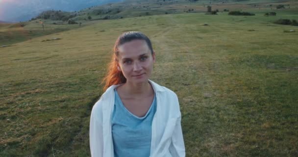 Donna felice turista si trova al campo di montagna al tramonto e sorride. Ritratto femminile. Vista sulle montagne e sul campo. — Video Stock