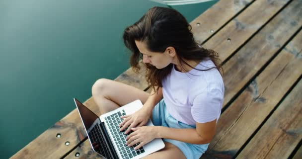 Gelukkig vrouw toerist zit op een houten pier en werkt aan een laptop, het typen van tekst. Uitzicht op het water. Uitzicht van bovenaf — Stockvideo