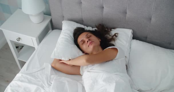 Молодая счастливая женщина просыпается. Женщина просыпается дома на белой кровати и поднимает руки. Женская улыбка — стоковое видео