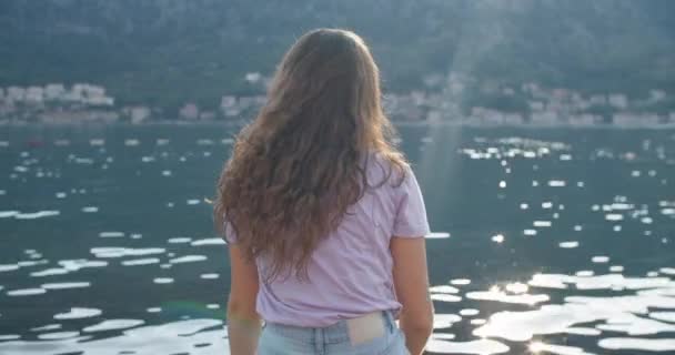 Šťastná brunetka sedí na dřevěném molu a má výhled na přírodu. Pohled na záliv a hory. Zadní pohled. — Stock video