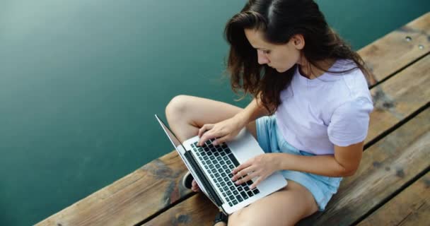 Donna felice turista è seduto su un molo di legno e lavorare su un computer portatile, digitando testo. Vista dell'acqua. Vista dall'alto — Video Stock