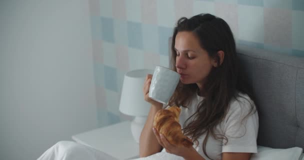 Mujer morena joven se despierta temprano en la mañana y desayuna con un croissant mientras está sentada en la cama. — Vídeo de stock