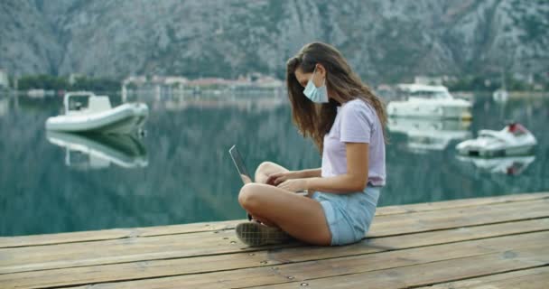 Vrouw toerist met beschermend masker zit op een houten pier en werkt aan een laptop, het typen van tekst. Uitzicht op het water. Uitzicht van bovenaf — Stockvideo