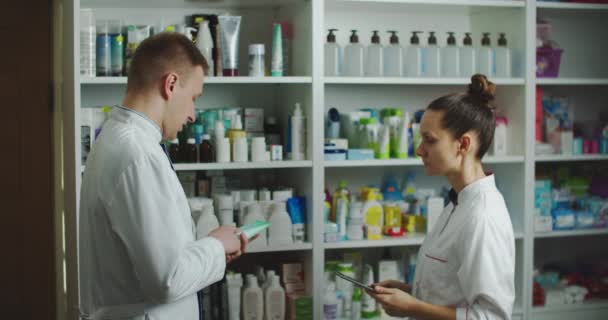 Dos farmacias hombre y mujer están en farmacia probando medicamentos en una tableta. Gran cheque o inventario — Vídeos de Stock