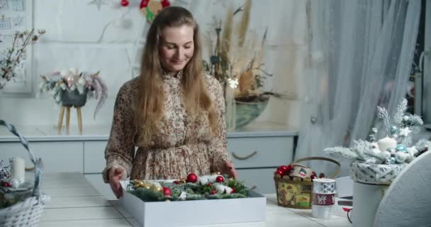 Μια γυναίκα πωλητής συσκευάζει ένα όμορφο στεφάνι Χριστουγέννων σε ένα κουτί προς πώληση. Χριστουγεννιάτικο δώρο — Αρχείο Βίντεο