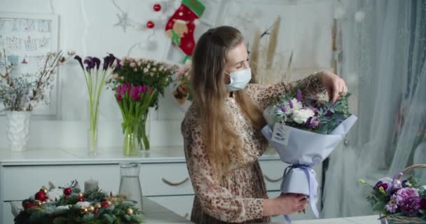 Bir çiçekçide tıbbi maske takan bir kadın satıcı satılık bir buket paketliyor. Bir salgın sırasında çiçek işi, virüs. — Stok video