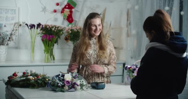 Una joven paga por la compra de un ramo de flores. Una hermosa chica sonriente está pagando con tarjeta de crédito por comprar flores frescas para un regalo a una joven vendedora de flores. — Vídeos de Stock