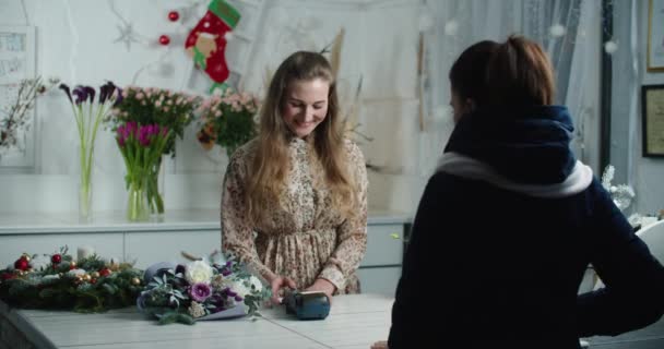 Ένα νεαρό κορίτσι πληρώνει για την αγορά ενός μπουκέτο λουλούδια. Ένα όμορφο χαμογελαστό κορίτσι πληρώνει με πιστωτική κάρτα για την αγορά νωπών λουλουδιών για ένα δώρο σε μια νεαρή γυναίκα ανθοπώλη. — Αρχείο Βίντεο