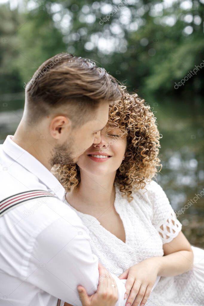 Happy husband kisses his wife with curly hair, sitting on the pier near the beautiful lake and forest. Close-up portrait, happy couple.