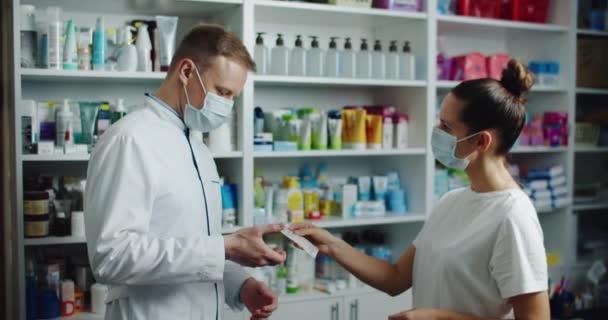 Egy gyógyszerész orvosi maszkban tanácsot ad egy vevőnek egy gyógyszertárban. Gyógyszerek vásárlása a vírus alatt. — Stock videók