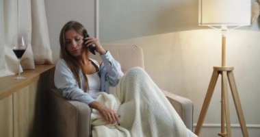 Mutlu esmer kadın beyaz bir battaniyenin altında koltuğa oturur, telefonda mesajlar okur ve kırmızı şarap içer. Evde akşam.